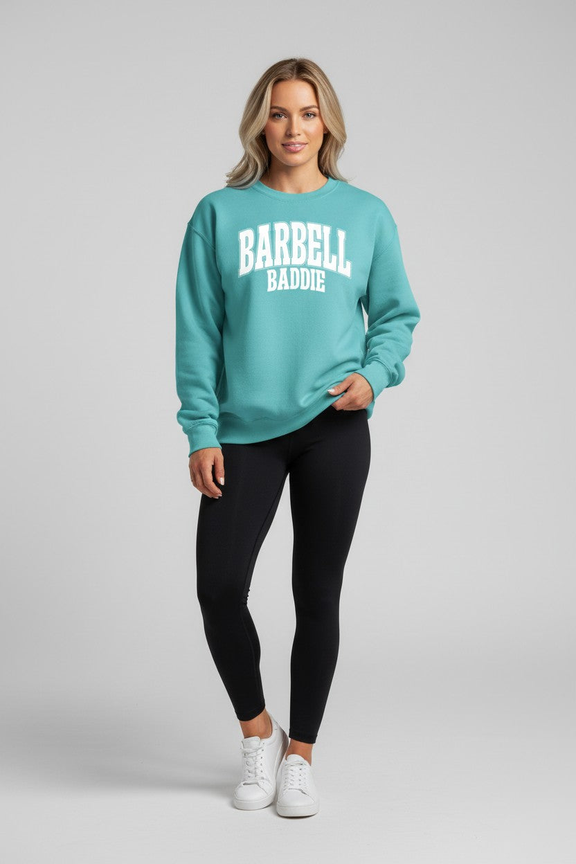 A woman modeling a teal crewneck sweatshirt with 'BARBELL BADDIE' printed in white across the chest, paired with black leggings and white sneakers, against a light gray studio background.