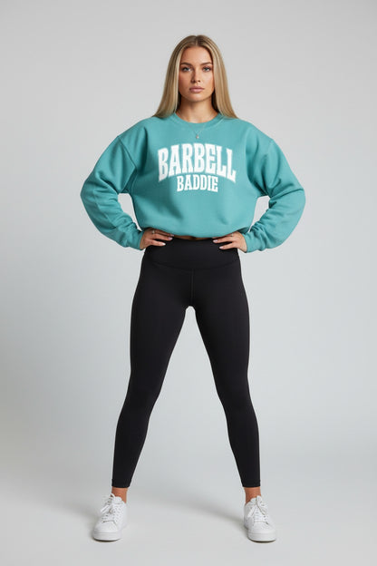 A woman modeling a teal sweatshirt with 'BARBELL BADDIE' printed in white on the front, paired with black leggings and white sneakers, against a light gray studio background.