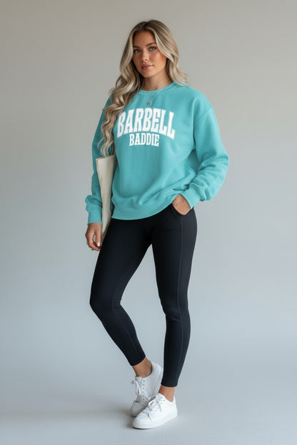 A woman modeling a teal sweatshirt with BARBELL BADDIE printed in white on the front, paired with black leggings and white sneakers, against a light gray studio background.