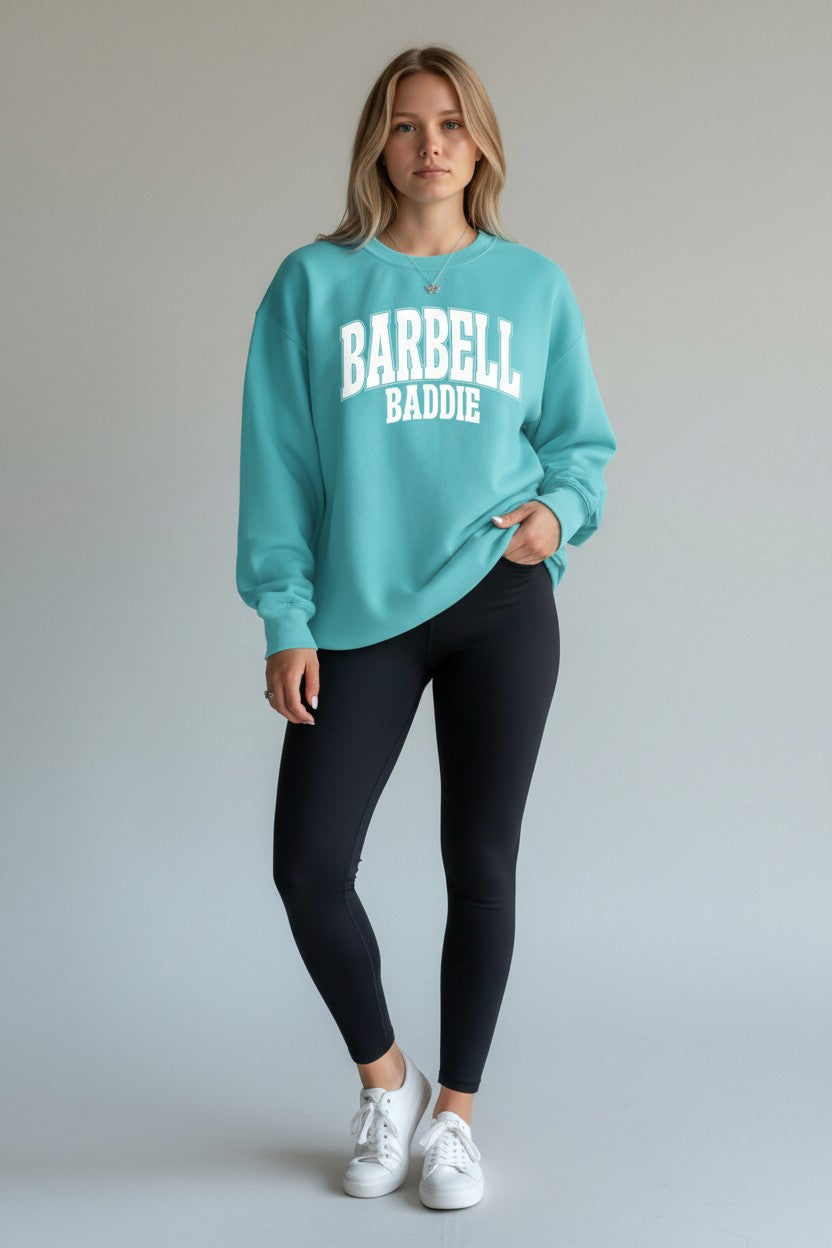 A woman modeling a turquoise crewneck sweatshirt with 'BARBELL BADDIE' printed in white across the chest, paired with black leggings and white sneakers, against a neutral gray studio background.