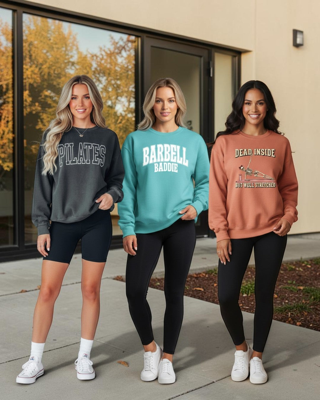 Three women modeling sweatshirts outdoors in front of a building with large windows. The sweatshirts are charcoal gray with 'PILATES' text, teal with 'BARBELL BADDIE' text, and terracotta orange with 'DEAD INSIDE' text, paired with black leggings and white sneakers.