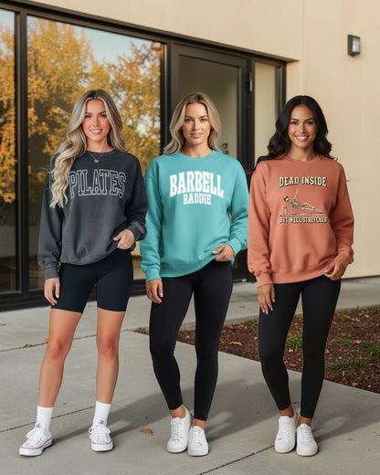 Three women modeling sweatshirts outdoors in front of a building with large windows. The sweatshirts are charcoal gray with 'PILATES' text, teal with 'BARBELL BADDIE' text, and terracotta orange with 'DEAD INSIDE' text, paired with black leggings and white sneakers.