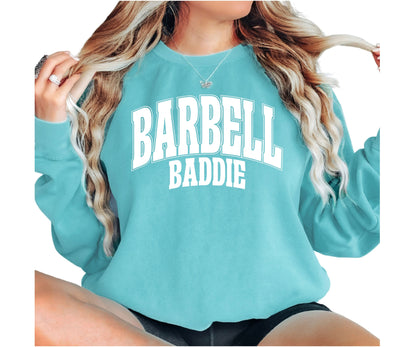 A woman wearing a turquoise crewneck sweatshirt with 'BARBELL BADDIE' printed in white varsity-style letters on the front, shown against a plain white background.