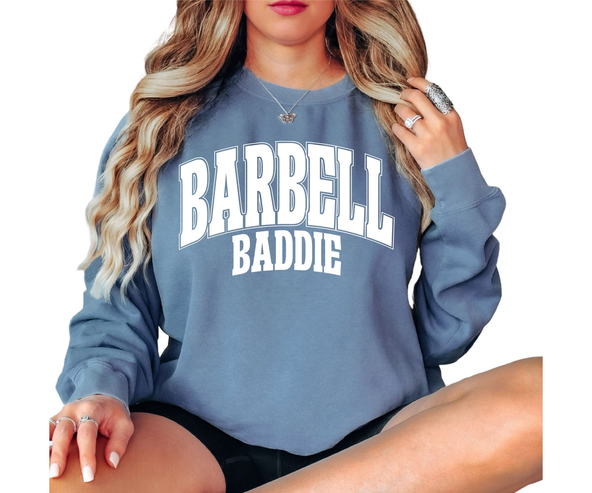 A woman wearing a slate blue sweatshirt with 'BARBELL BADDIE' printed in white varsity-style letters across the chest, shown against a plain white background.