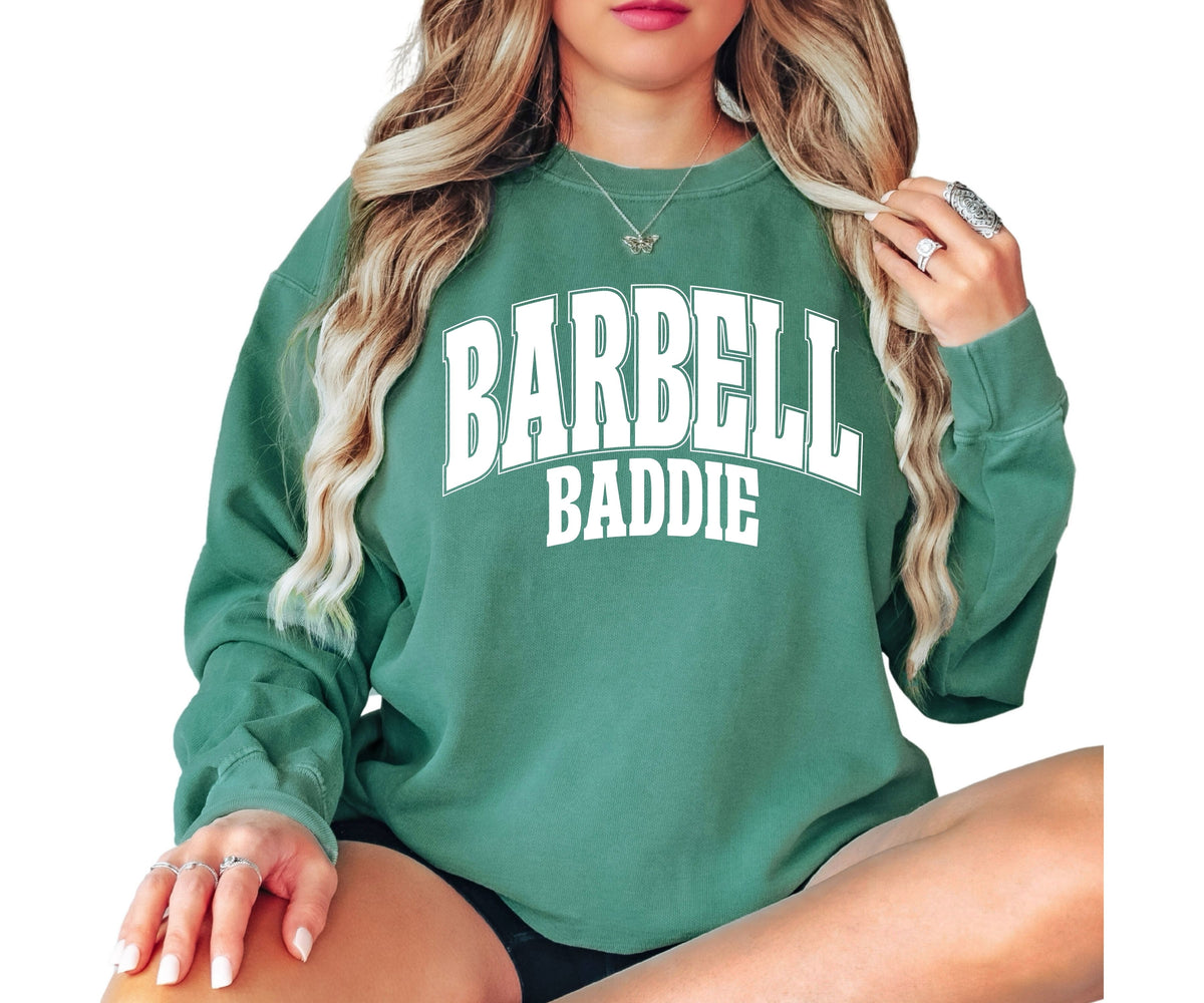 A woman wearing a green crewneck sweatshirt with 'BARBELL BADDIE' printed in white arched text on the front, shown against a plain white background.