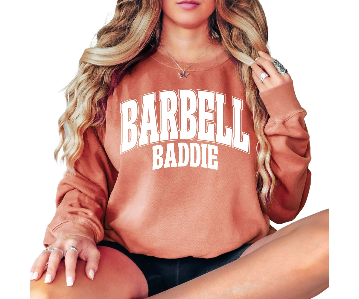 A woman wearing a terracotta-colored crewneck sweatshirt with 'BARBELL BADDIE' printed in white varsity lettering across the chest, posed against a solid white background.