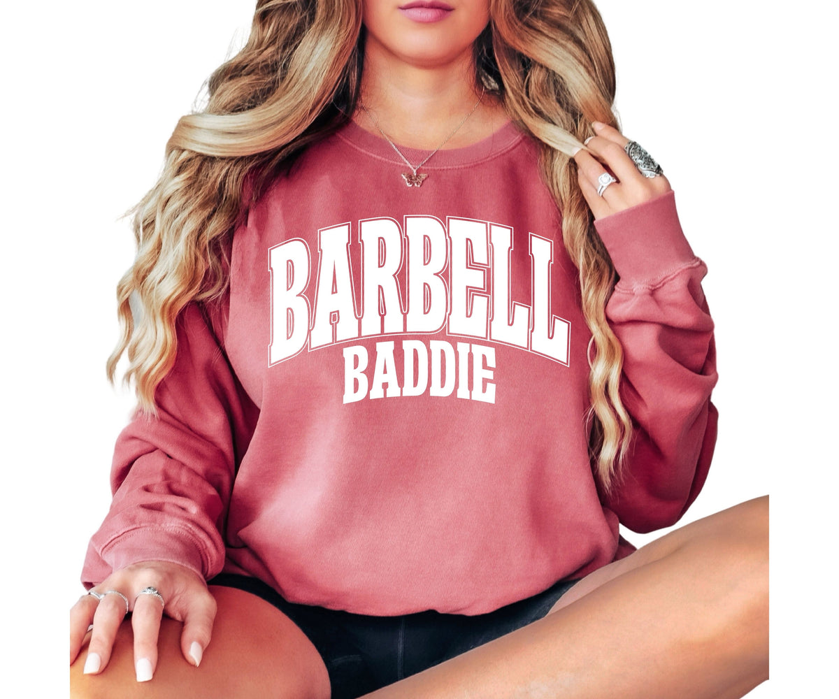 A woman wearing a dusty rose crewneck sweatshirt with 'BARBELL BADDIE' printed in white varsity lettering across the chest, shown against a plain white background.