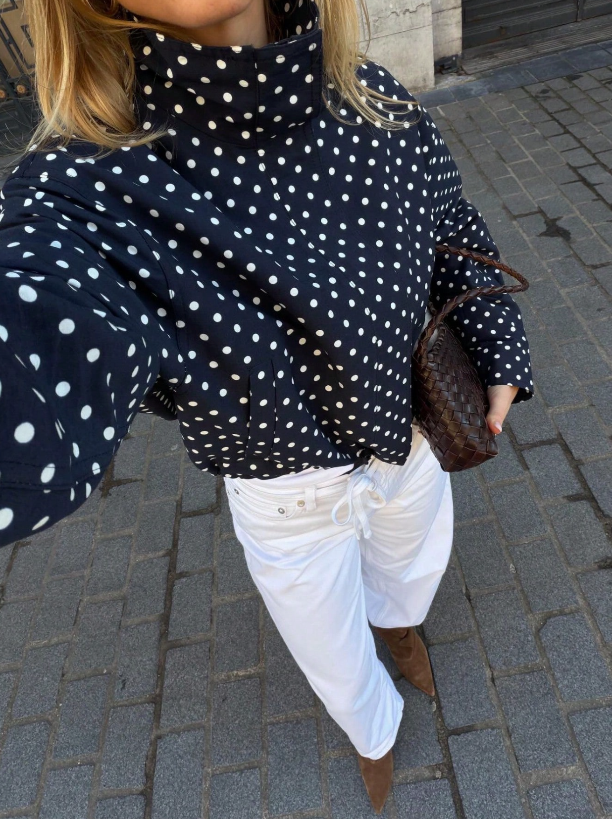 A woman takes a selfie wearing a navy blue long-sleeve turtleneck top with white polka dots, white drawstring pants, and brown ankle boots while carrying a brown woven handbag on a gray paved street.