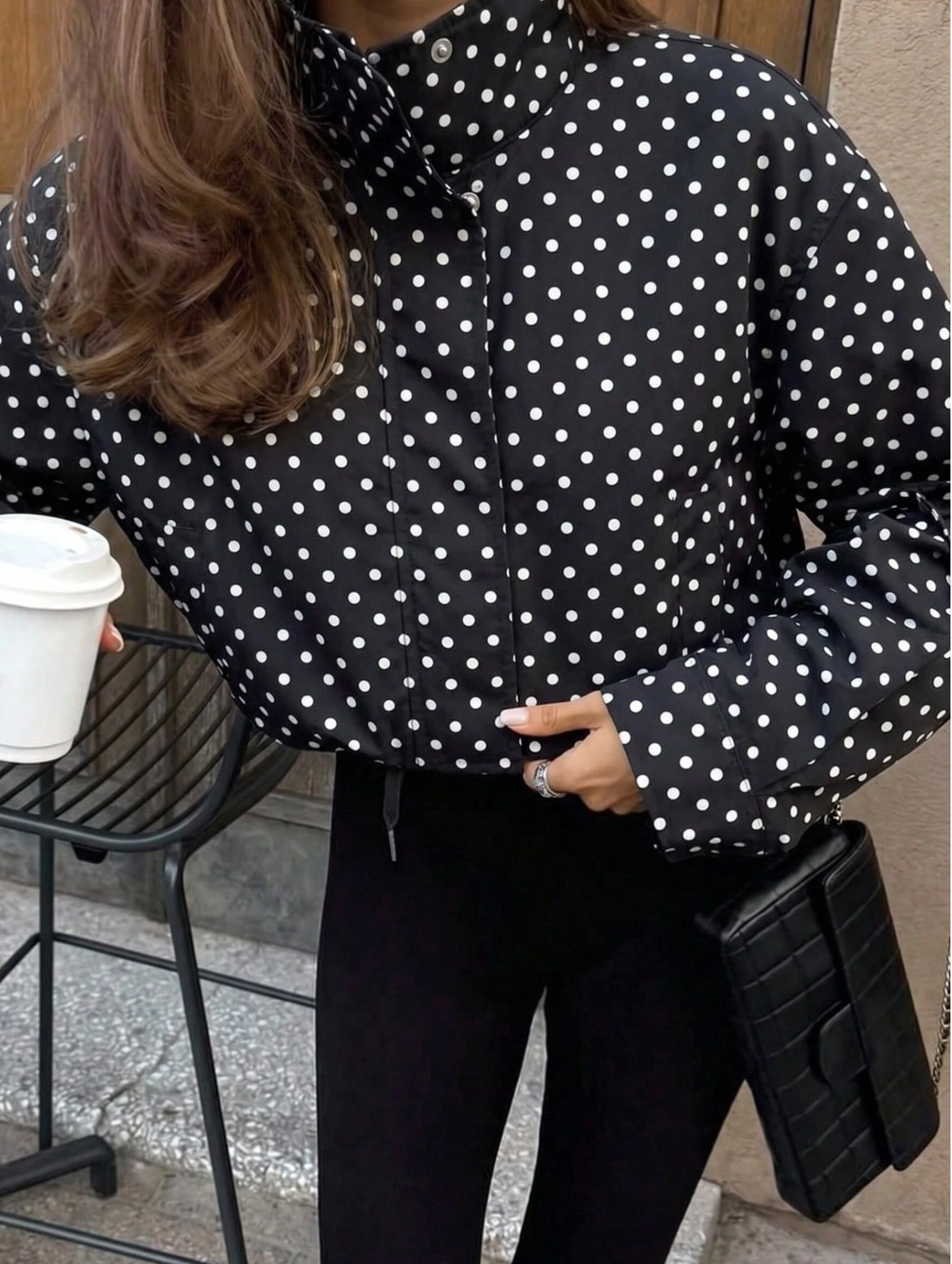 A person wearing a black puffer jacket with a white polka dot pattern, paired with black pants, holding a black clutch bag and standing next to a black metal chair.