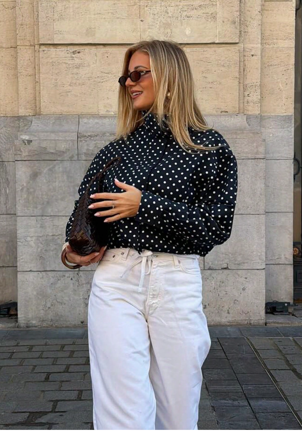 A woman wearing a black long-sleeve blouse with white polka dots and white drawstring trousers, standing outdoors against a stone wall.