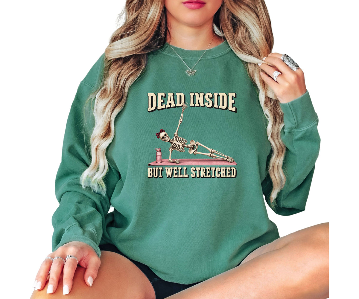 A woman wearing a green crewneck sweatshirt with a graphic of a skeleton stretching and the text 'DEAD INSIDE BUT WELL STRETCHED'.