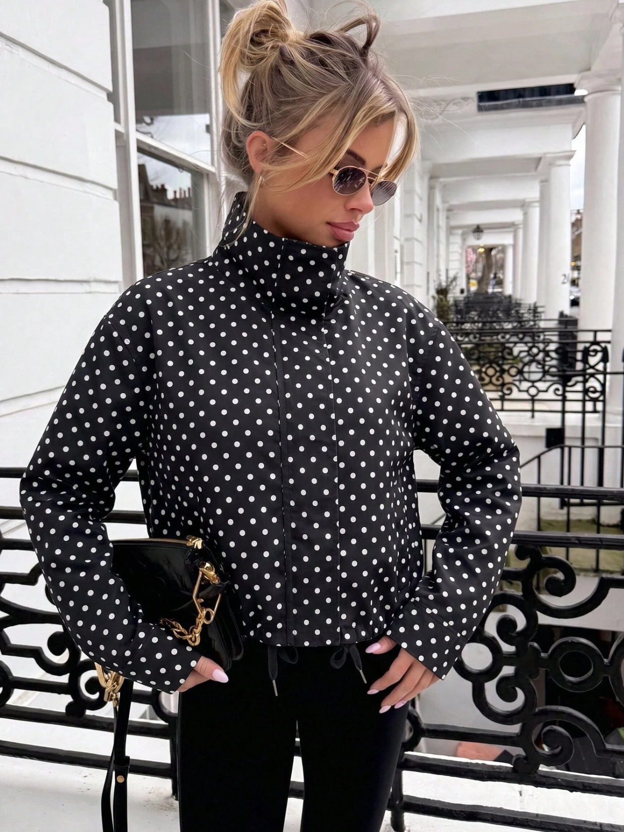 A woman wearing a black long-sleeve mock-neck top with a white polka dot pattern, styled with black pants and sunglasses, holding a black handbag with gold hardware.