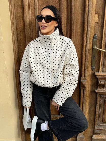 A woman wearing a white long-sleeve mock-neck blouse with black polka dots, paired with black jeans, black sneakers, and black sunglasses, posing in front of a rustic wooden door.