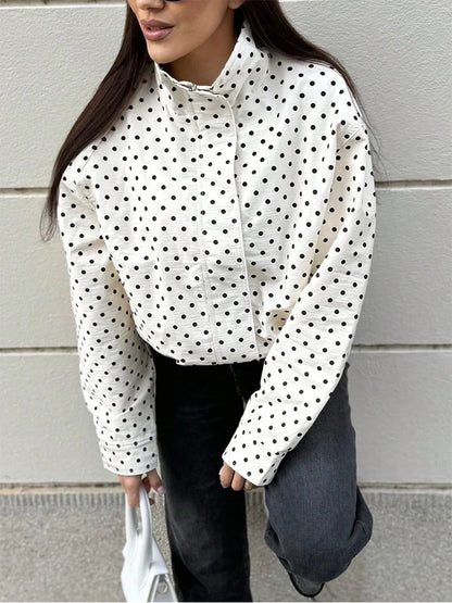 A woman wearing a white long-sleeve blouse with a black polka dot pattern, paired with dark gray jeans, standing against a light gray paneled wall.