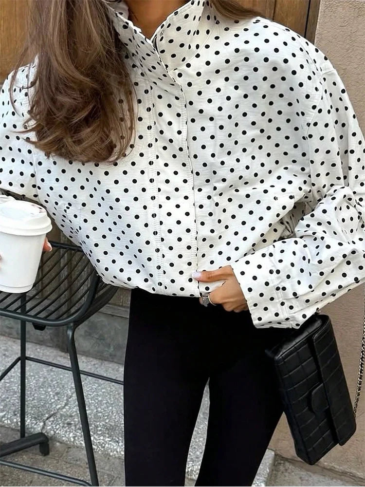 A woman wearing a white long-sleeve button-up blouse with black polka dots, paired with black pants and holding a black quilted clutch bag.