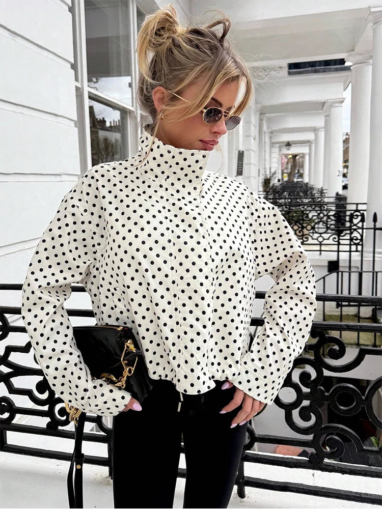 A woman wearing a white long-sleeve mock neck blouse with black polka dots, paired with black pants and a black handbag, standing on a white porch with black wrought-iron railings.