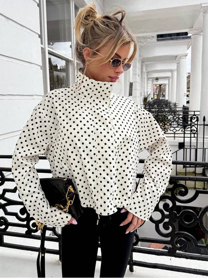 A woman wearing a white long-sleeve mock neck blouse with black polka dots, paired with black pants and a black handbag, standing on a white porch with black wrought-iron railings.