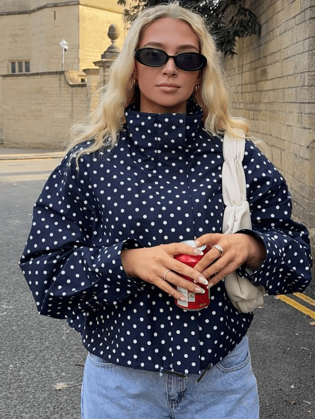 A woman wearing a navy blue and white polka dot mock neck top, black sunglasses, and holding a red can, standing outdoors against a brick wall.