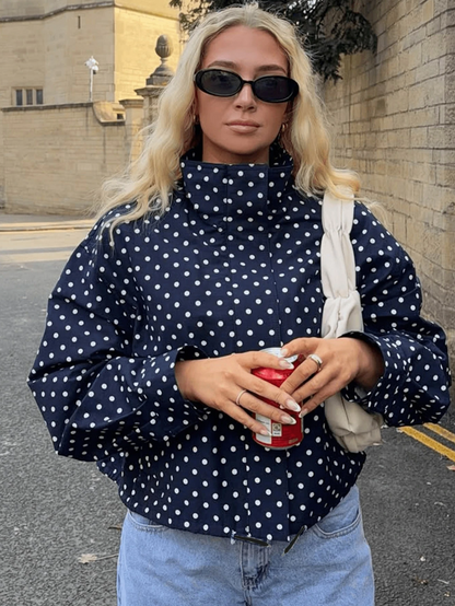 A woman wearing a navy blue and white polka dot mock neck top, black sunglasses, and holding a red can, standing outdoors against a brick wall.